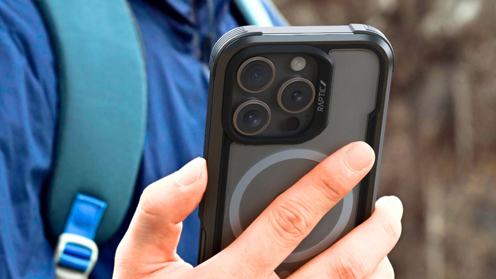 A person wearing a blue jacket and backpack is holding a smartphone with a rugged case, showing its triple-lens camera setup.
