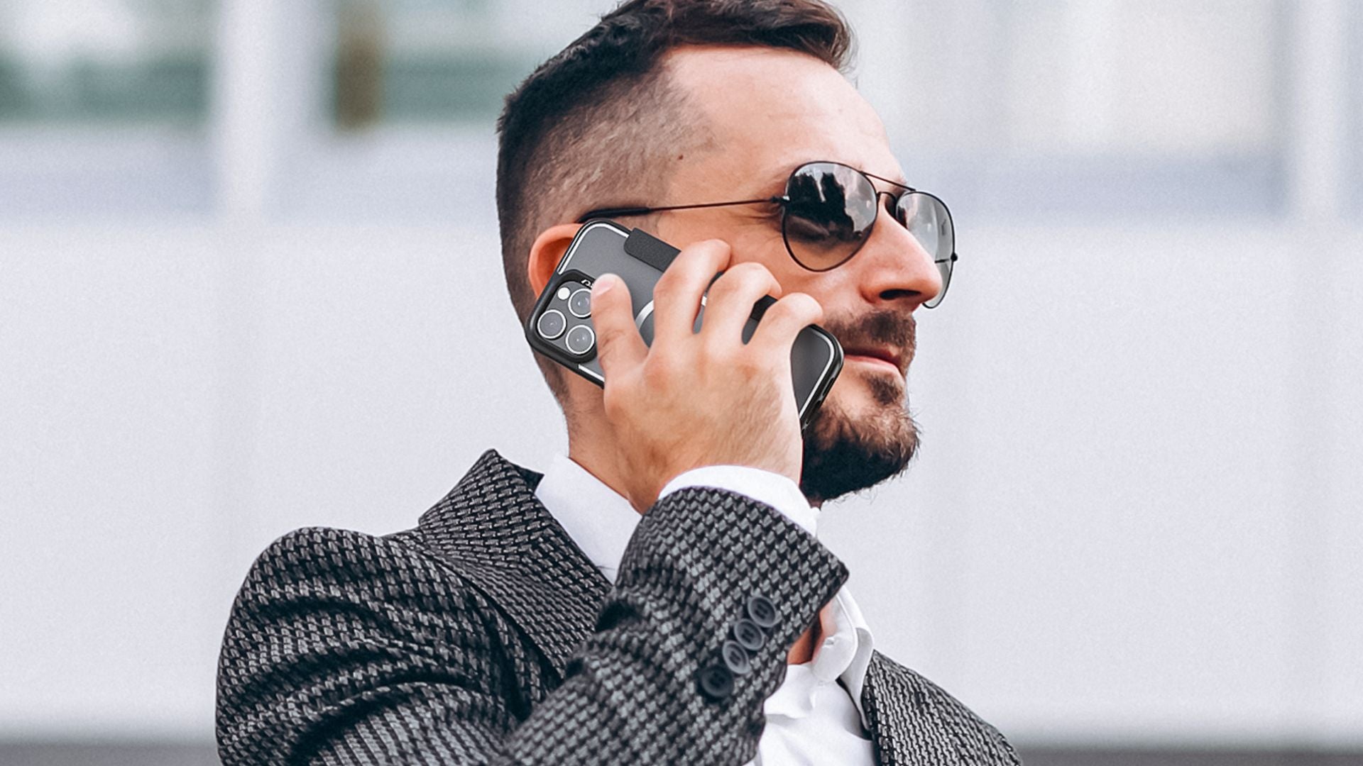 Hands holding a smartphone horizontally in an outdoor setting, showing the back of the phone with a case featuring a visible circular design around the Apple logo and a triple camera setup.