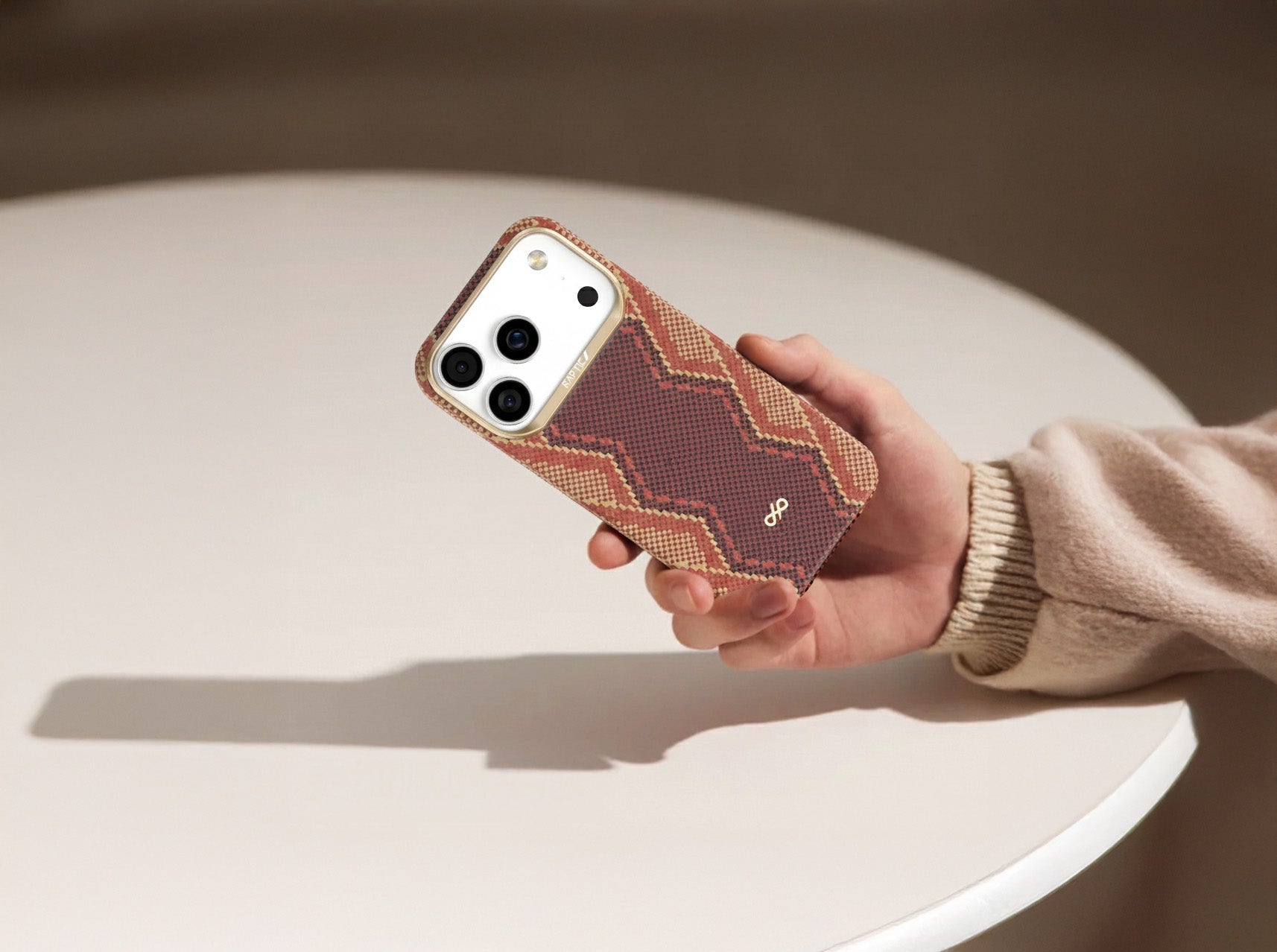 A hand in a beige sweater holds a smartphone with a brown, tan, and red zigzag-patterned case over a round white table. The phone's three camera lenses are clearly visible.