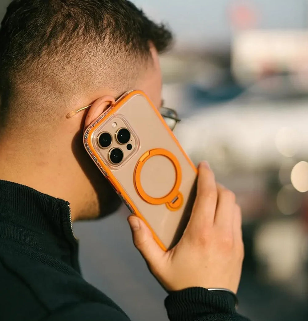 A person with short hair, wearing glasses and a dark jacket, is holding an iPhone 16 Pro Max with a Raptic AirJoy Beads transparent case featuring air bumpers and a bright orange ring. They seem engrossed in conversation outdoors, as the blurred background suggests.
