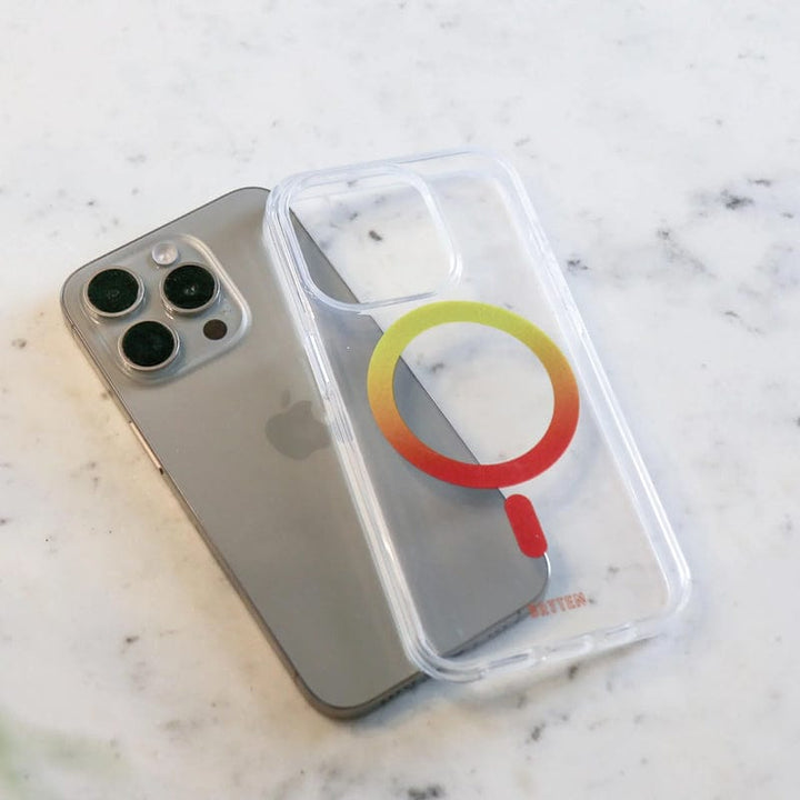 A silver iPhone with three cameras rests on a white marble surface next to the Bryten by Raptic iPhone 15 MagFx MagSafe Case, which features a clear design with a colored circle, showcasing its wireless charging compatibility and drop protection.