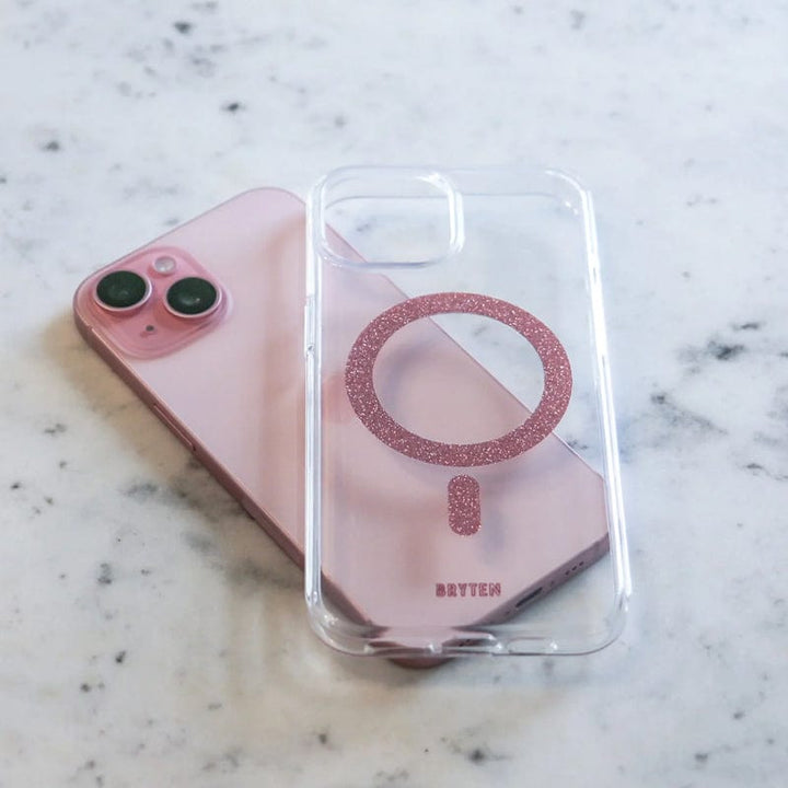 A pink smartphone lies on a marble surface next to a Bryten by Raptic iPhone 15 MagFx MagSafe Case, which features a clear design with a glittery pink ring and supports wireless charging.