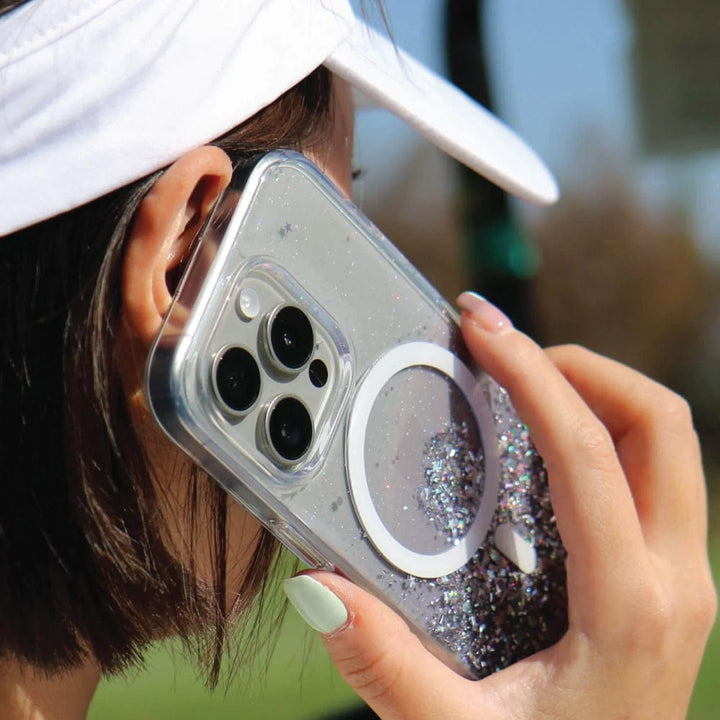Person holding an iPhone 15 Pro Max with a Bryten by Raptic iPhone 15 Series Starburst MagSafe Case to their ear. The phone has three camera lenses and a ring on the back for holding. The glittery, transparent case features seamless MagSafe integration, while the person is wearing a white visor.