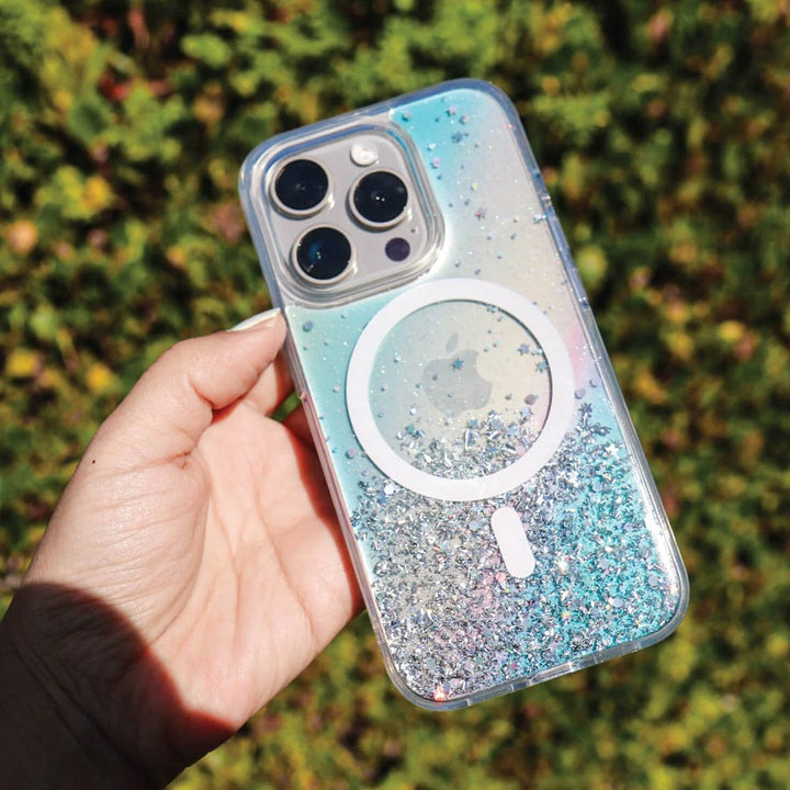 A hand holds an iPhone encased in a Bryten by Raptic iPhone 15 Series Starburst MagSafe Case, which features sparkling glitter and a circular design on the back. This Bryten case offers Seamless MagSafe Integration for effortless charging. The background of greenery accentuates its reflective iridescent design.