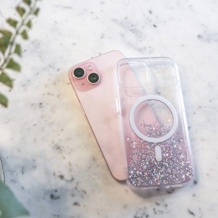 A pink smartphone with dual cameras lies on a marble surface next to a Bryten by Raptic iPhone 15 Series Starburst MagSafe Case filled with glitter and a circular grip, boasting seamless MagSafe integration.