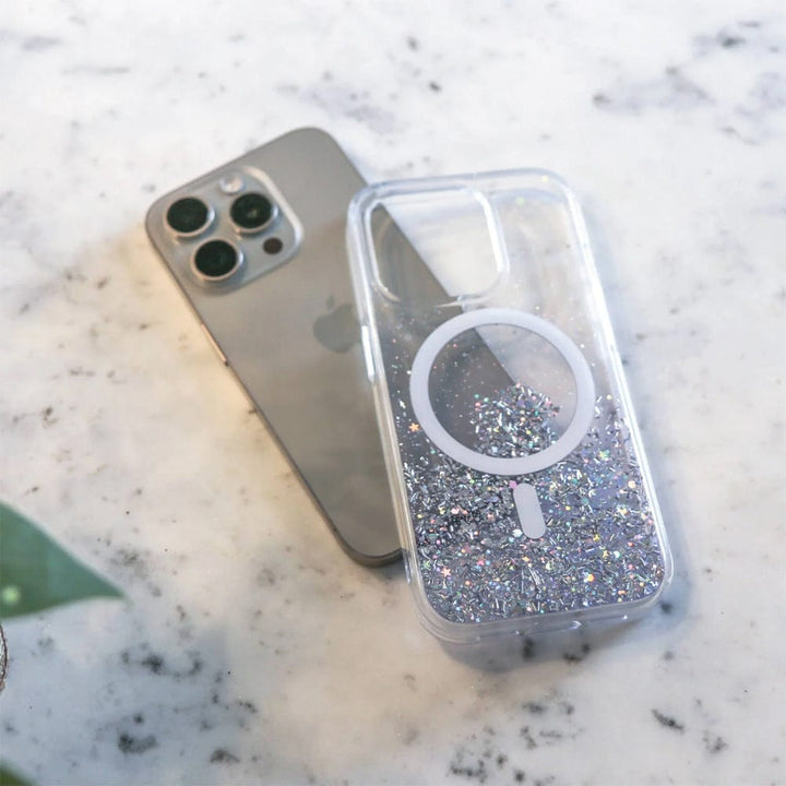 A gold smartphone with three cameras is placed on a marble surface next to a Bryten by Raptic iPhone 15 Series Starburst MagSafe Case, which is clear, filled with silver glitter, and features a white magnetic ring to offer seamless MagSafe integration.