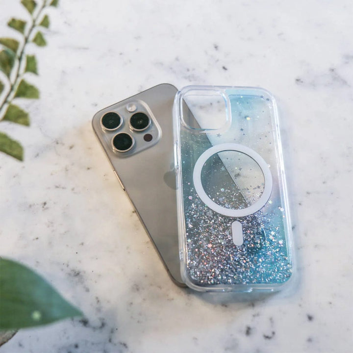 An iPhone with a triple camera setup lies next to the Bryten by Raptic iPhone 15 Series Starburst MagSafe Case, which is glittery and transparent, on a marble surface, with a green plant in the background.