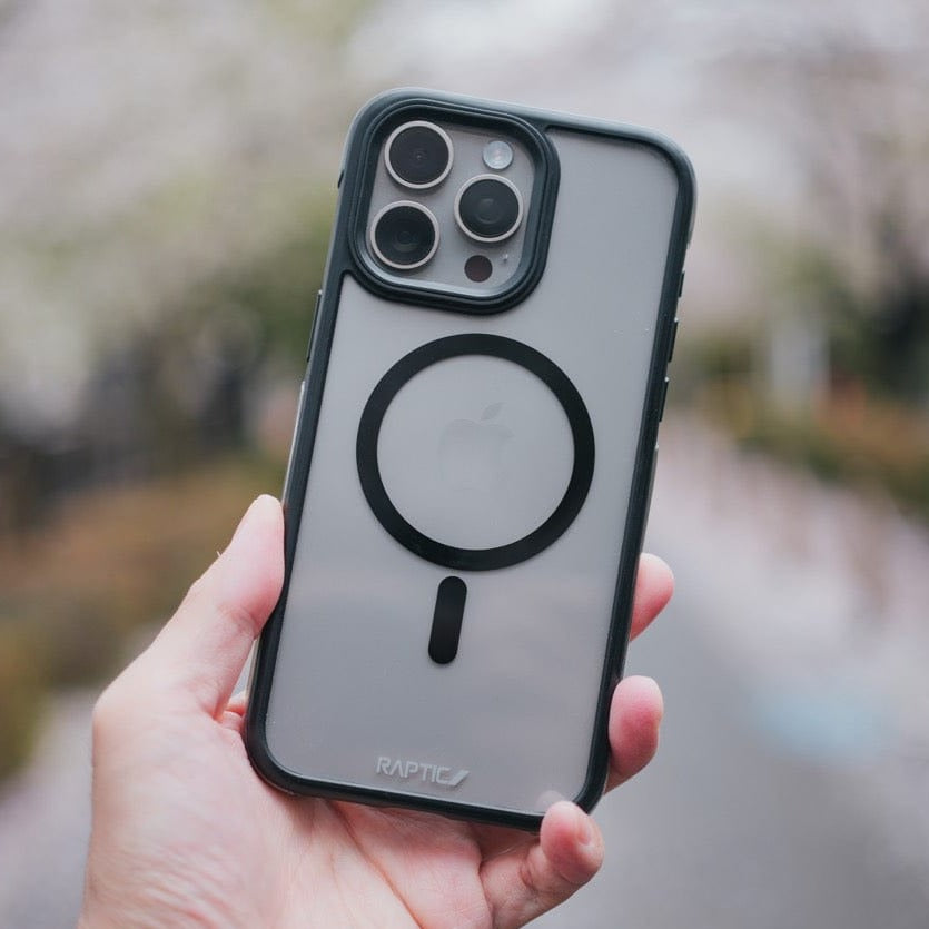 A hand holding an iPhone 15 Air Plus with a black Raptic case featuring Military Spec drop protection, displaying three camera lenses and an apple logo, against a blurred background of blossoming trees.