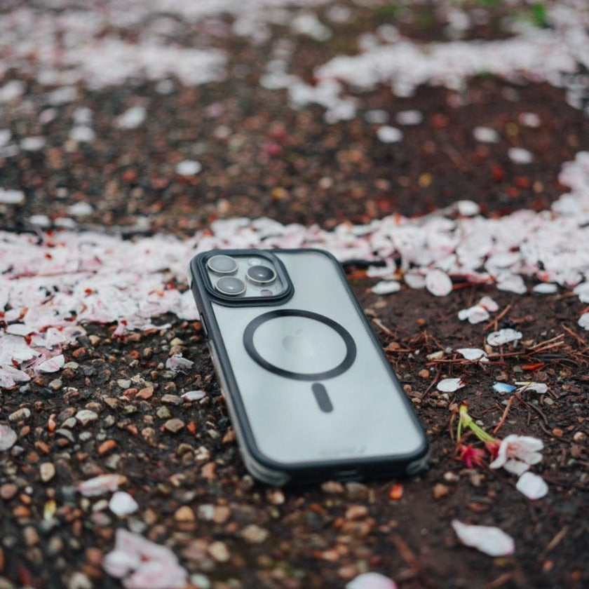 The Raptic Air+ Slim iPhone 15 Series in a black case with Military Spec drop protection lies face up on a wet, gravelly surface with scattered pink and white flower petals around it.