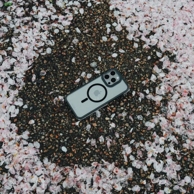 A Raptic Air+ Slim iPhone 15 Series with Military Spec drop protection case lies on a ground covered with scattered pink flower petals.