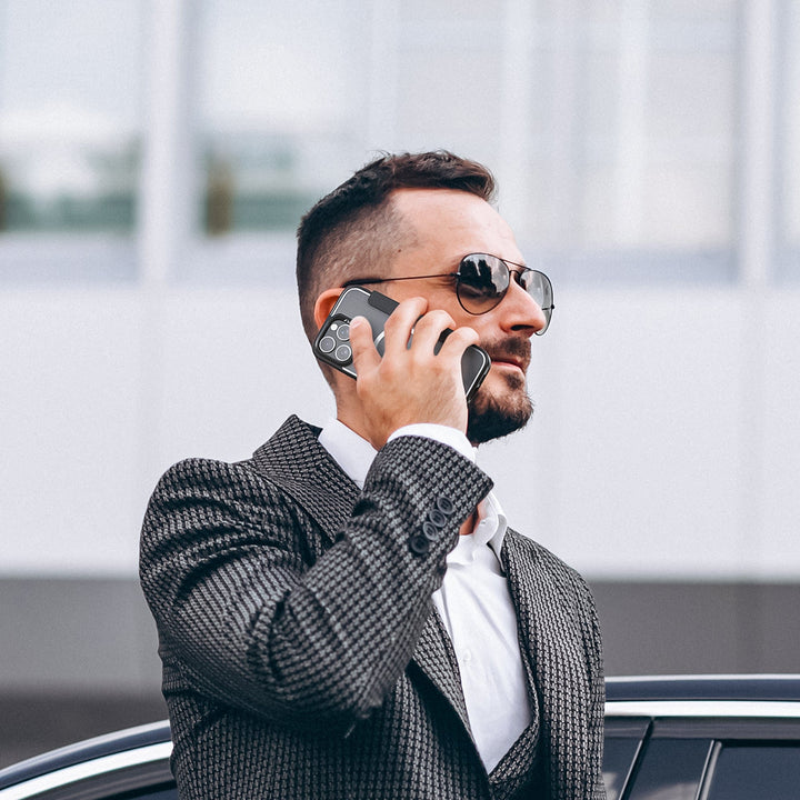 A man with short dark hair, a beard, and sunglasses is standing outside, holding a Raptic MagSafe Urban Folio Case with Card Slot for iPhone 16 to his ear. He is wearing a textured gray suit with a white dress shirt. A portion of a car and a modern building with large windows are visible in the background.