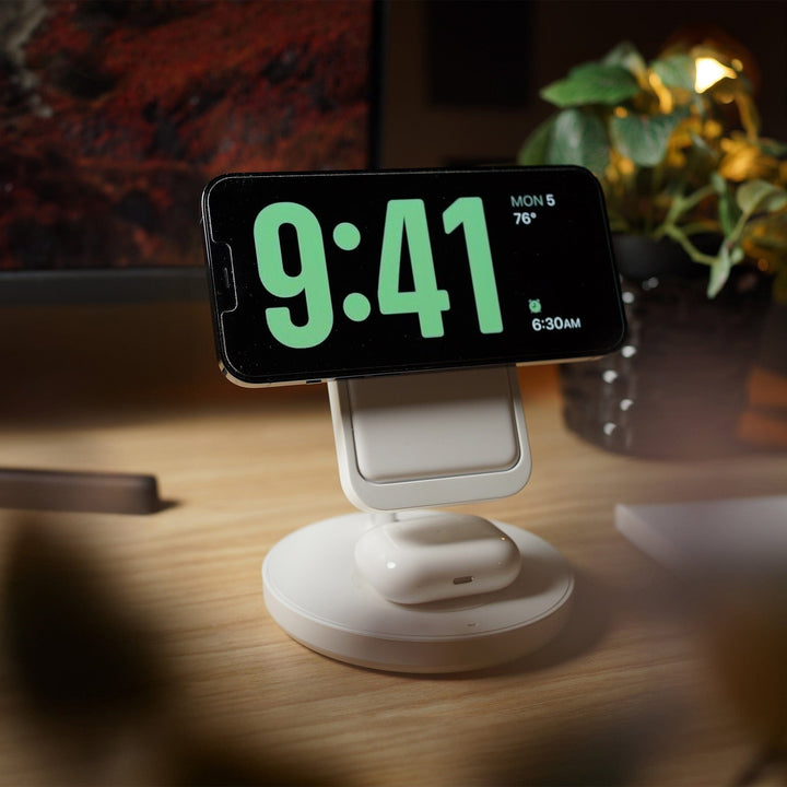 An Urban 6-in-1 MagSafe M6 Wireless Charger Station is showcasing a smartphone displaying the time "9:41" and the date "Mon 5" docked on its white wireless charging stand. Below the phone, a pair of white wireless earbuds are also charging on the same station. The setup, featuring an iPhone-compatible magnetic case, is placed on a wooden desk with a computer monitor in the background and a potted plant to the right.