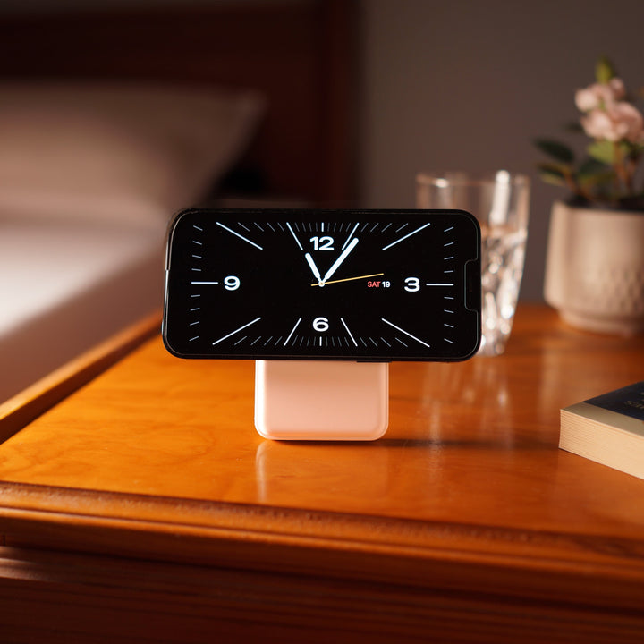 A smartphone displaying a digital clock rests horizontally on an Urban 6-in-1 MagSafe M6 Wireless Charger Station atop a white stand on a wooden bedside table. The table also holds a glass of water, a potted plant, and a book. Behind it, the neatly made bed with white linens is softly illuminated in the background.