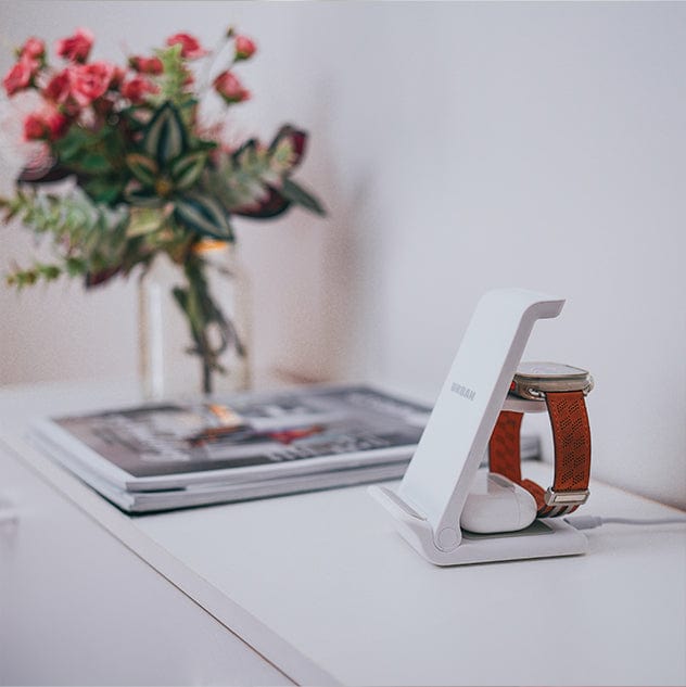 An Urban Travel S 3 in 1 Wireless Charger by Urban holds a smartwatch and wireless earbuds on a white desk. In the background, there is a vase with flowers and a stack of magazines.