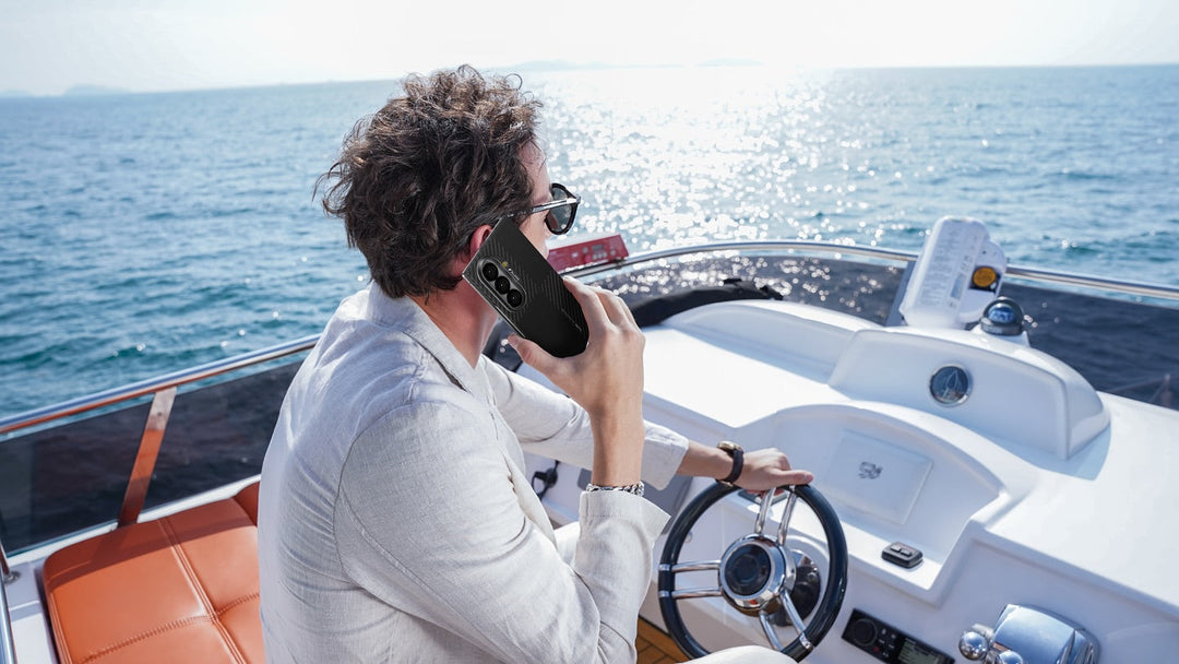 A man in a light suit sits at the helm of a boat, steering with one hand and talking on a smartphone. The boat is on open water under a sunny, clear sky.