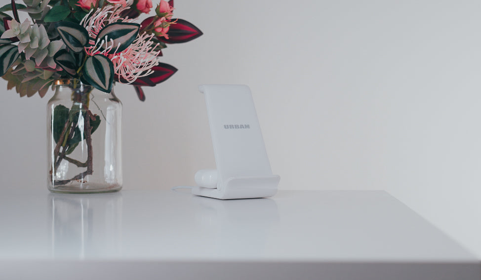 A white phone stand labeled "URBAN" sits on a white surface next to a glass vase with pink and green flowers.