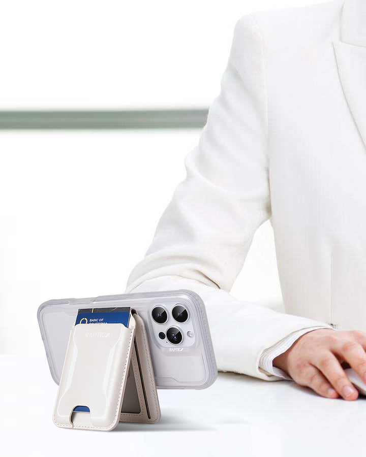 A person in a white suit sits at a table. Up front, the Raptic iPhone 17 Magnetic Wallet - Raptic Air Wallet with a white cardholder stands upright, showing a bank card and an iPhone with three rear cameras against a minimal, blurred background.