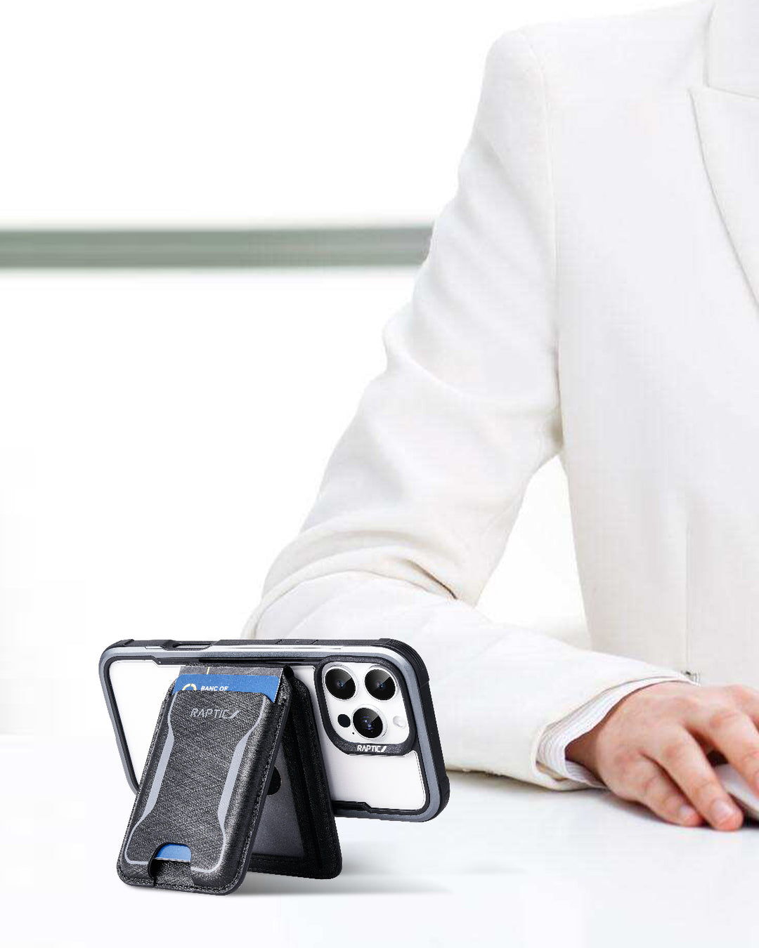 A person in a white suit sits at a desk, showing only their arm and hand. In front is the Raptic iPhone 17 Magnetic Wallet - Air Wallet in black with a blue accent, propped on its built-in MagSafe stand and displaying triple camera lenses.