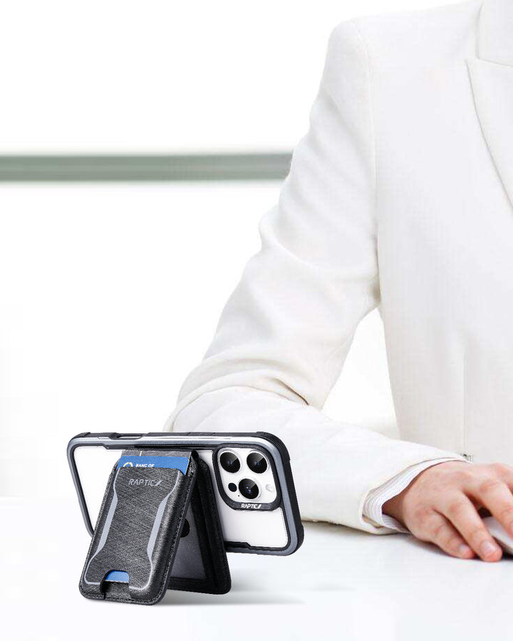 A person in a white suit sits at a desk, showing only their arm and hand. In front is the Raptic iPhone 17 Magnetic Wallet - Air Wallet in black with a blue accent, propped on its built-in MagSafe stand and displaying triple camera lenses.