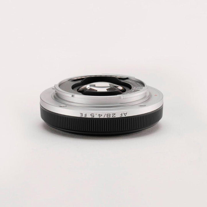 A silver and black Viltrox 28mm f/4.5 FE wide-angle lens for Sony full-frame cameras lies flat on a white surface, showcasing its compact build, ribbed black grip, visible glass elements, and minimalistic background.