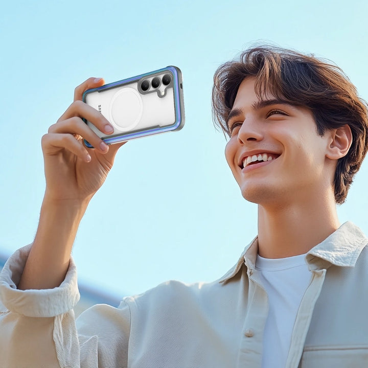 A young brown-haired man is outdoors, smiling and holding a Samsung Galaxy S25 with a Raptic Shield case. He's dressed in a light jacket and white shirt against a bright blue sky.