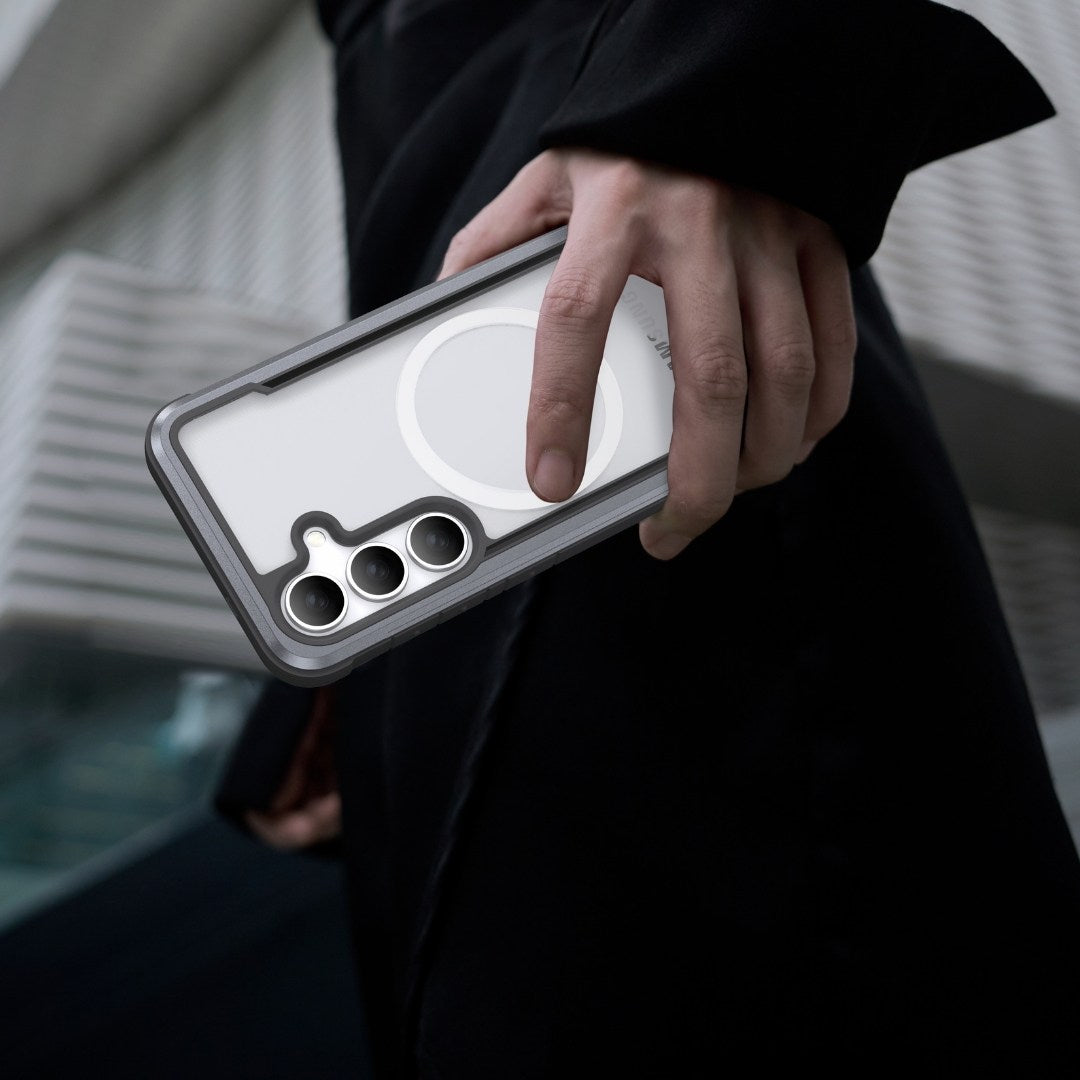 A person in a black coat holds a Raptic Shield Samsung S25 case with a frosted white finish in their right hand. The case offers military-grade protection and shock absorption, showcasing a circular design with three visible camera lenses against a blurred background of diagonal architectural lines.