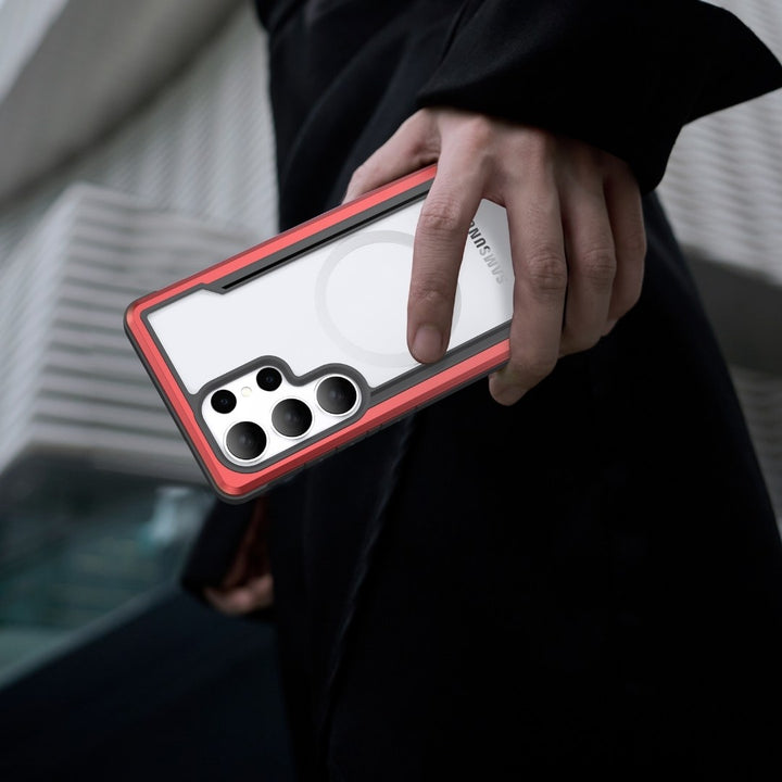 A person holds a red and white Raptic Samsung S25 Shield Case, featuring military-grade protection and a unique circular design. The phone with multiple camera lenses is set against a geometric building, while the individual in black clothing prominently displays their hand.