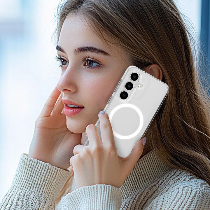 A woman with long brown hair holds a white smartphone featuring a Raptic Crystal MagSafe case for the Samsung S25 Series. The transparent case displays a circular design indicating MagSafe compatibility. She wears a cream-colored knit sweater and looks slightly aside, set against a soft-focus background.