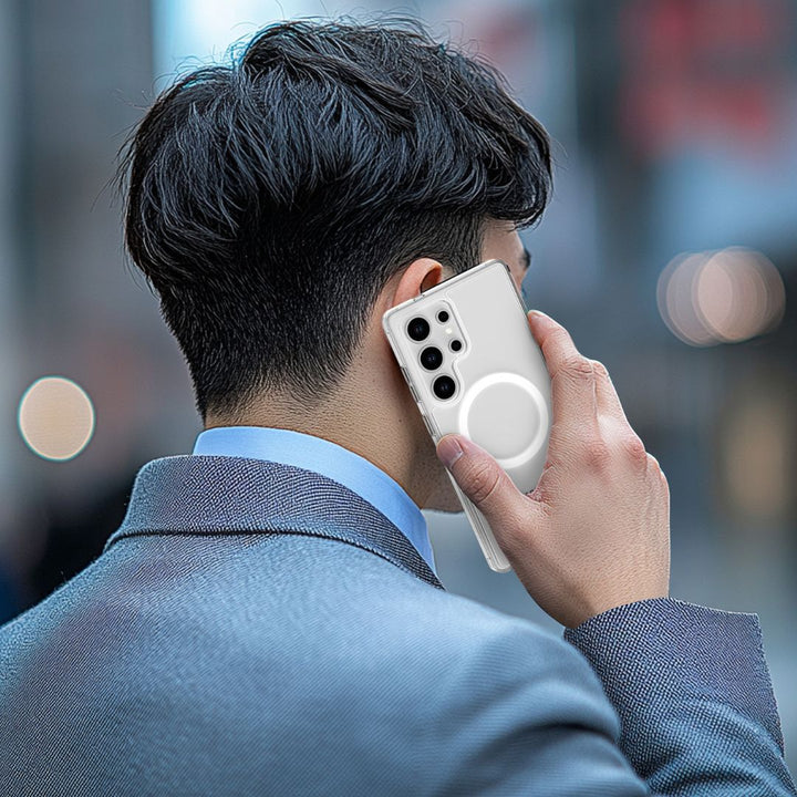 A person in a light grey suit holds a phone to their ear, protected by a Raptic Crystal MagSafe case for the Samsung S25 Series featuring a central circular design. The blurred background suggests an urban setting, with the individual's short dark hair visible but face not shown.