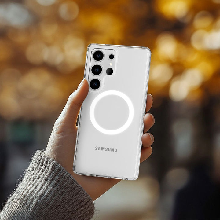 A hand holding a Samsung S25 with a Raptic Crystal MagSafe case reveals its glowing light and multiple lenses. The case provides drop protection, while the blurred orange and brown autumn leaves in the background create a warm atmosphere.