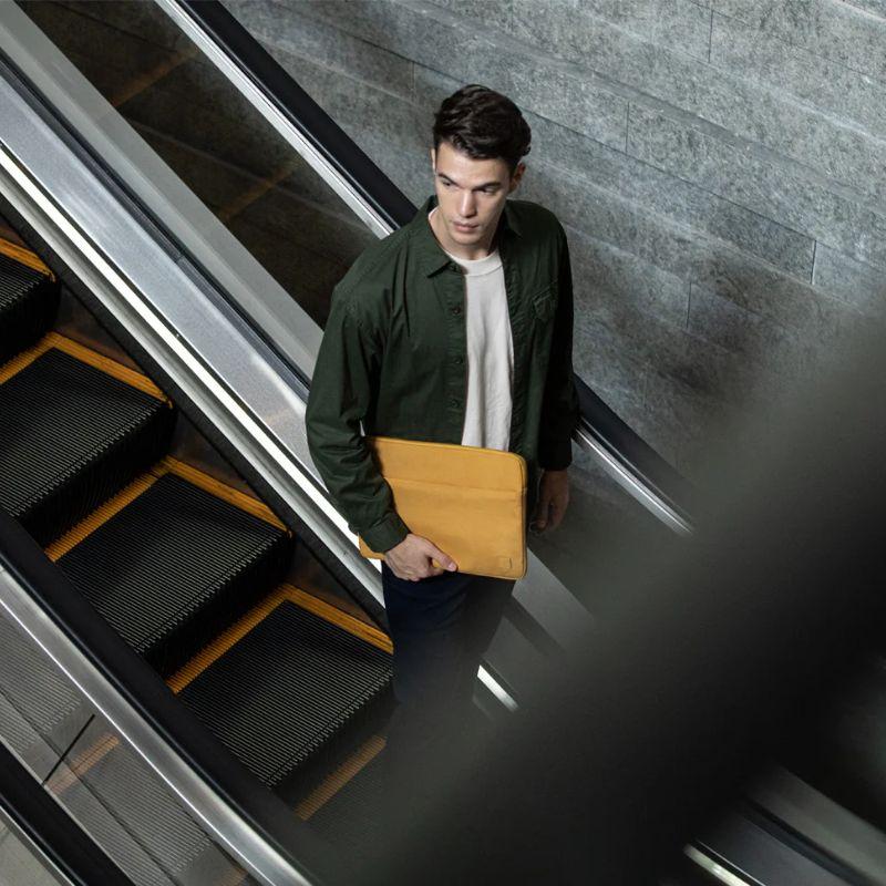 A young man with dark hair and a green jacket over a white shirt stands on an escalator, holding the UNIQ Vienna Shockproof Laptop Carry Sleeve for MacBook Pro 16-inch. The gray tiled wall is in the background, seen from above through railings.