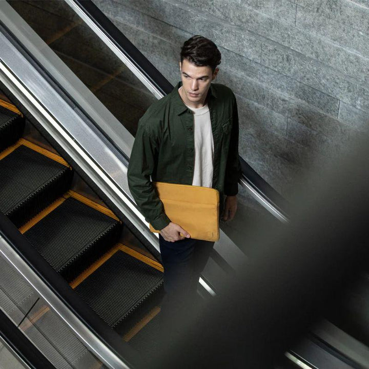 A young man with dark hair and a green jacket over a white shirt stands on an escalator, holding the UNIQ Vienna Shockproof Laptop Carry Sleeve for MacBook Pro 16-inch. The gray tiled wall is in the background, seen from above through railings.
