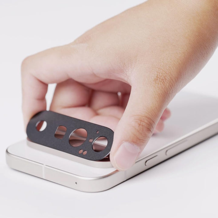 A hand holds the MobileTechnica Google Pixel 10 5G Camera Lens Protector above the rear camera of a silver phone, ready to apply. The phone, with a white surface and an aplus carousel container beside it, awaits the anti-scratch tempered glass.