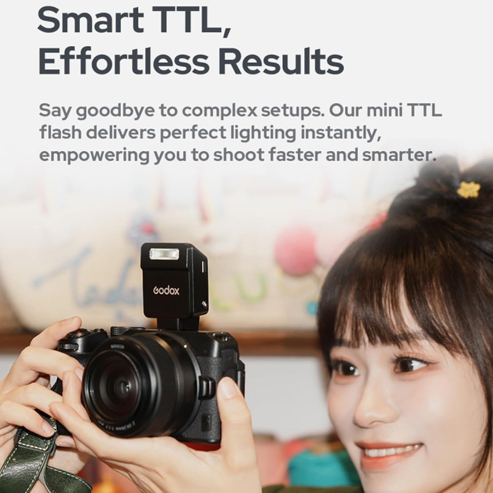 A woman with long dark hair and a colorful clip smiles, holding a camera fitted with the MobileTechnica Godox iT20-F TTL Mini Flash Speedlight for Fujifilm cameras. Soft lighting and a blurred background showcase this compact, premium accessory.