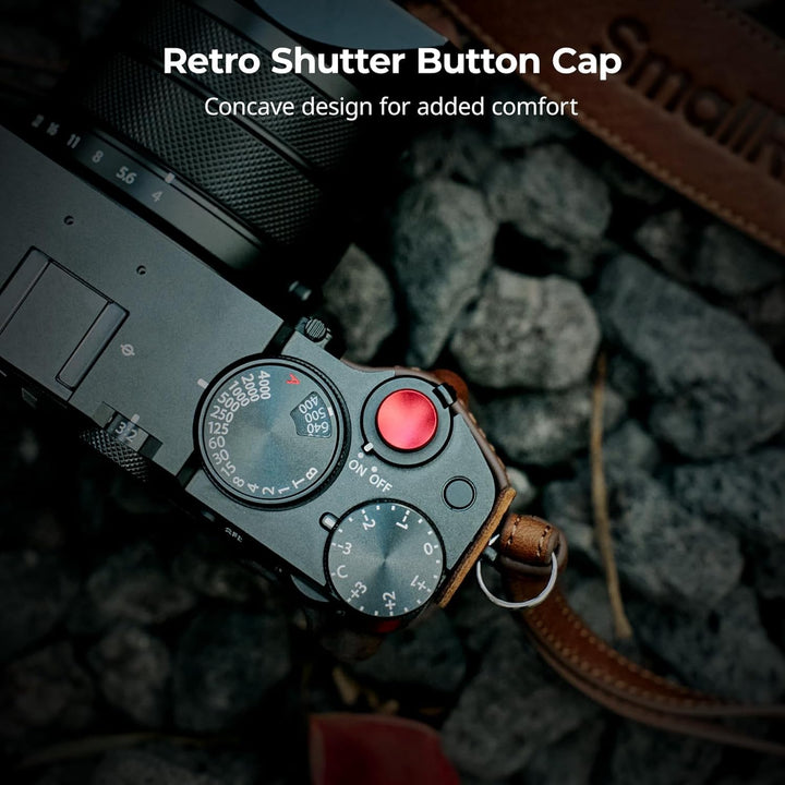 A close-up of a FUJIFILM GFX100RF with the SmallRig GFX100RF Leather Case Kit: leather case, black thumb pad, shoulder strap, and concave red shutter button cap for comfort. Camera rests on gray rocks. Text: “Retro Shutter Button Cap—Concave comfort.”.