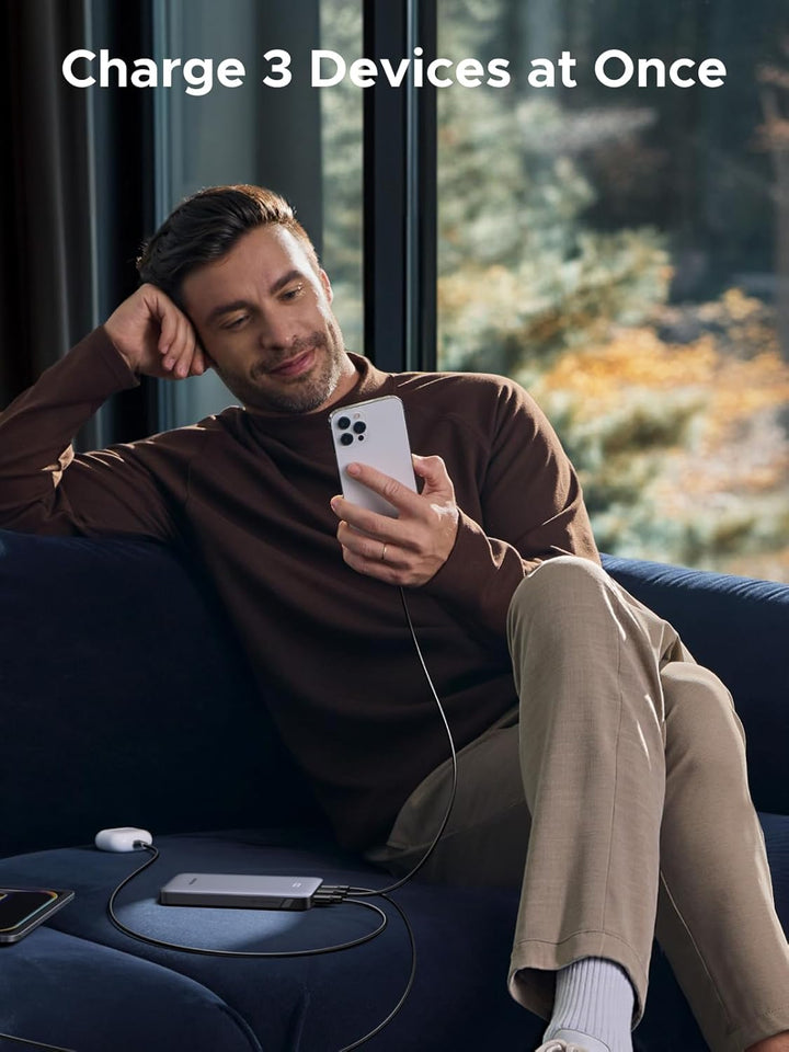 A man smiles on a blue sofa by a window, using his phone. Next to him is the UGREEN Slim Power Bank 10000mAh 30W PD, charging three devices at once. White text above says, "Charge 3 Devices at Once.