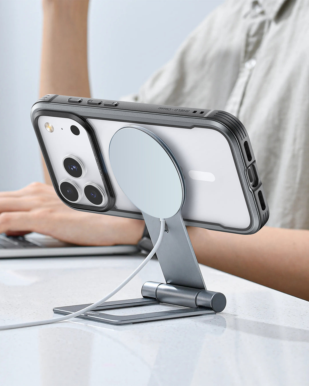 A person types on a laptop while an iPhone 17 Raptic Shield Glass Bundle Pack by Raptic sits horizontally on a silver adjustable stand, charging via cable, all resting on a white glossy tabletop.