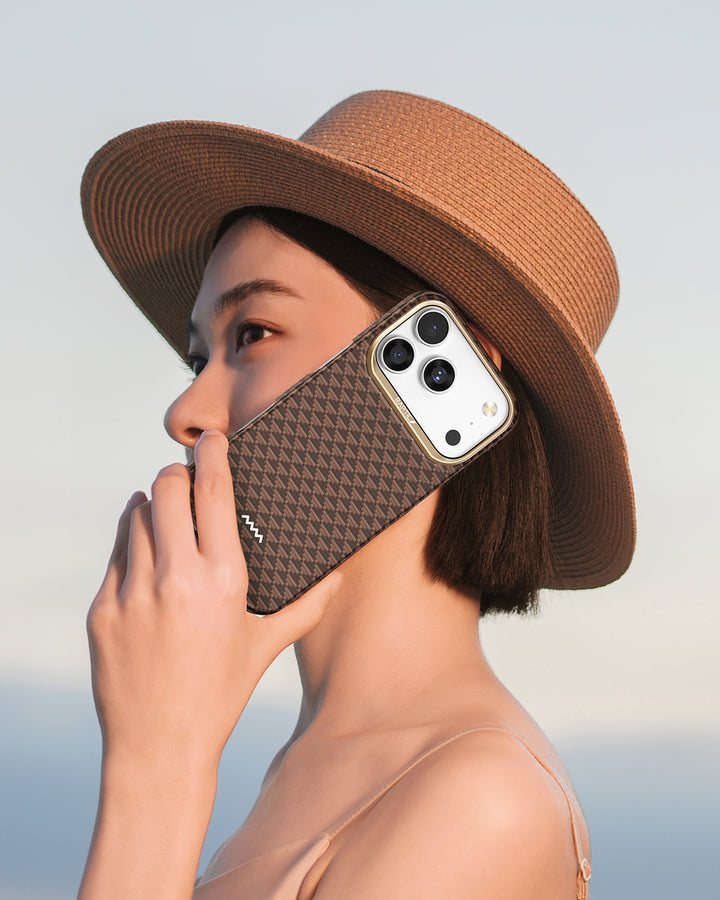 A woman with short dark hair in a straw hat and sleeveless top holds an iPhone 17 clad in the ultra-slim Raptic Skin-Origin Aramid Fibre Case to her ear, poised as if calling, against a soft sky backdrop.