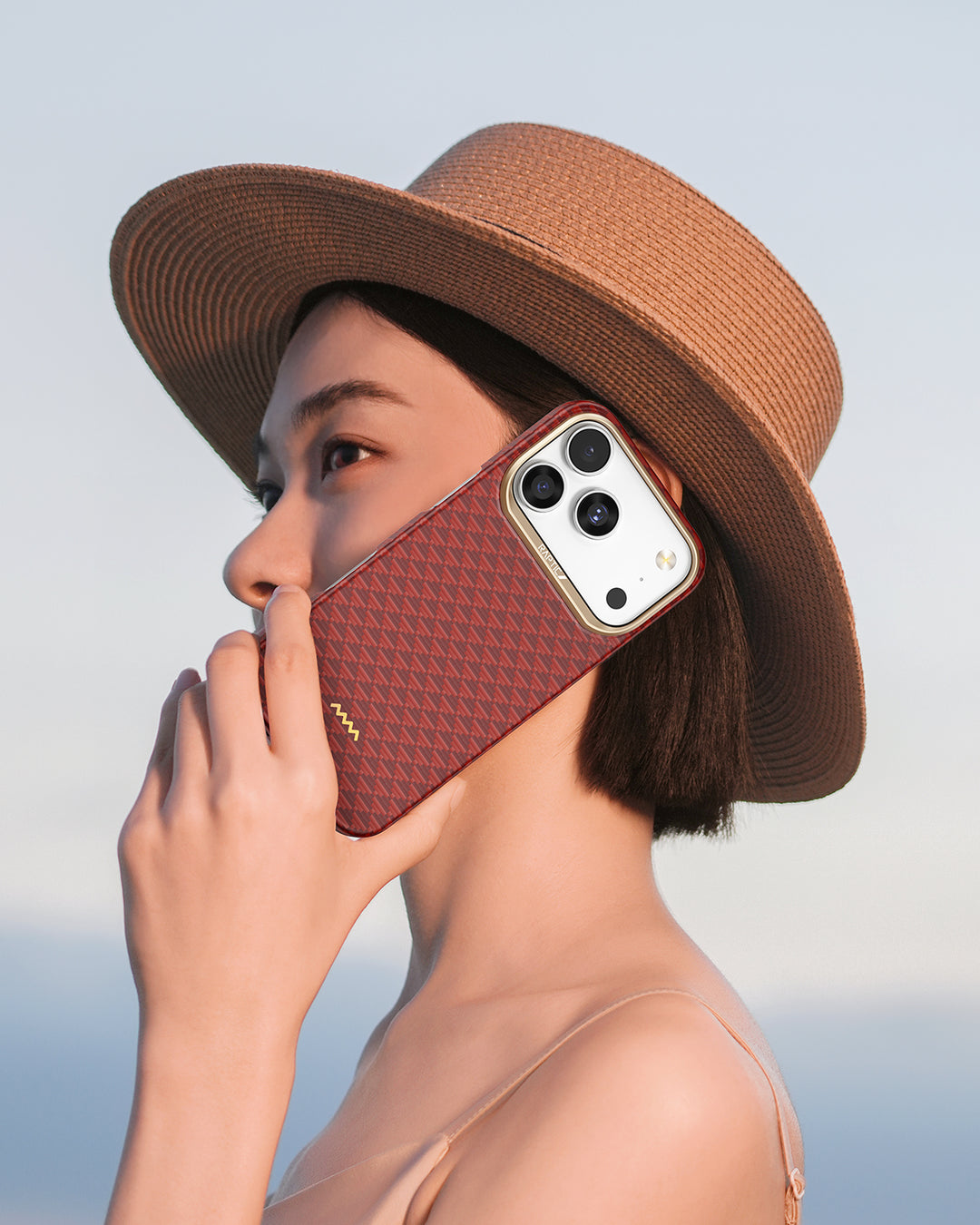 A woman with short dark hair, wearing a wide-brimmed brown hat and thin-strapped top, holds a Raptic iPhone 17 Aramid Fibre Case - Skin-Origin to her ear, gazing into the distance against a pale sky background.