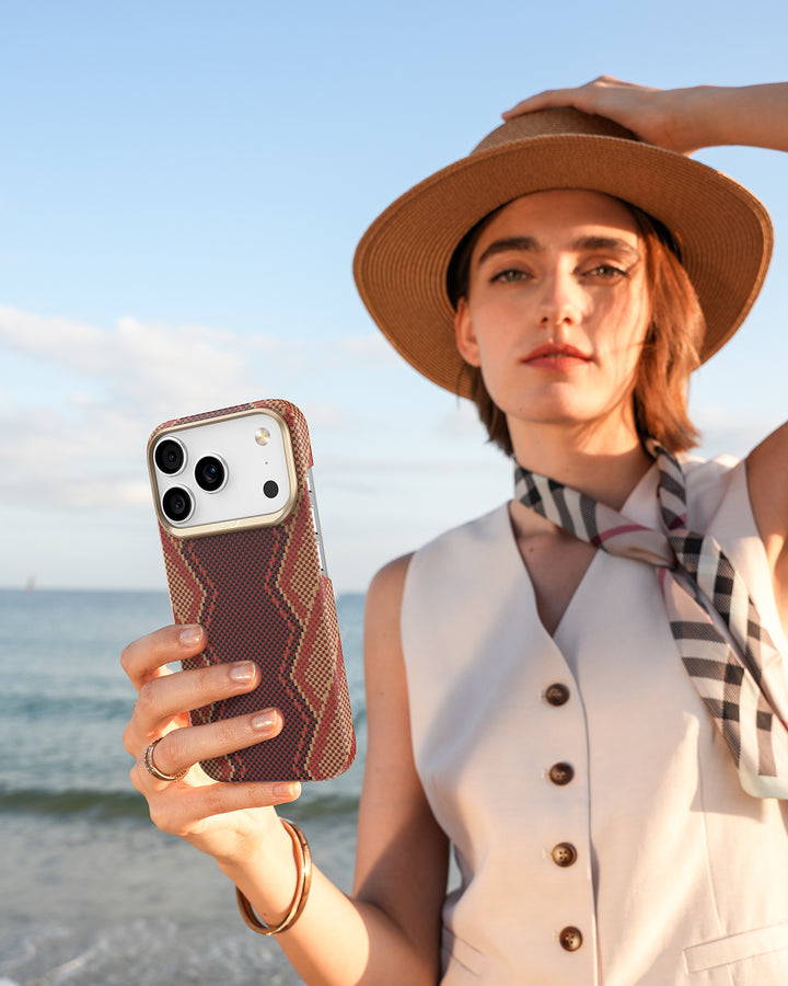A woman in a beige vest, patterned scarf, and hat stands by the seaside, holding her iPhone 17 in a Raptic Skin-Origin Aramid Fibre Case. Sunlight lights her face as she looks at the camera with ocean waves behind her.