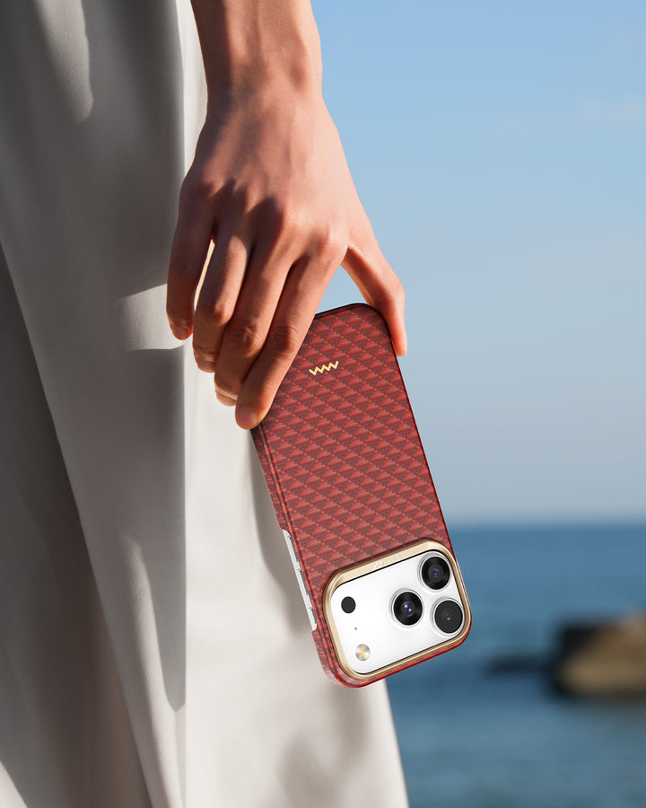 A person holds a Raptic iPhone 17 Aramid Fibre Case – Skin-Origin, featuring a red geometric pattern and small gold logo. The blurred beachy backdrop hints at a sunny day, while the user wears light-colored clothing.