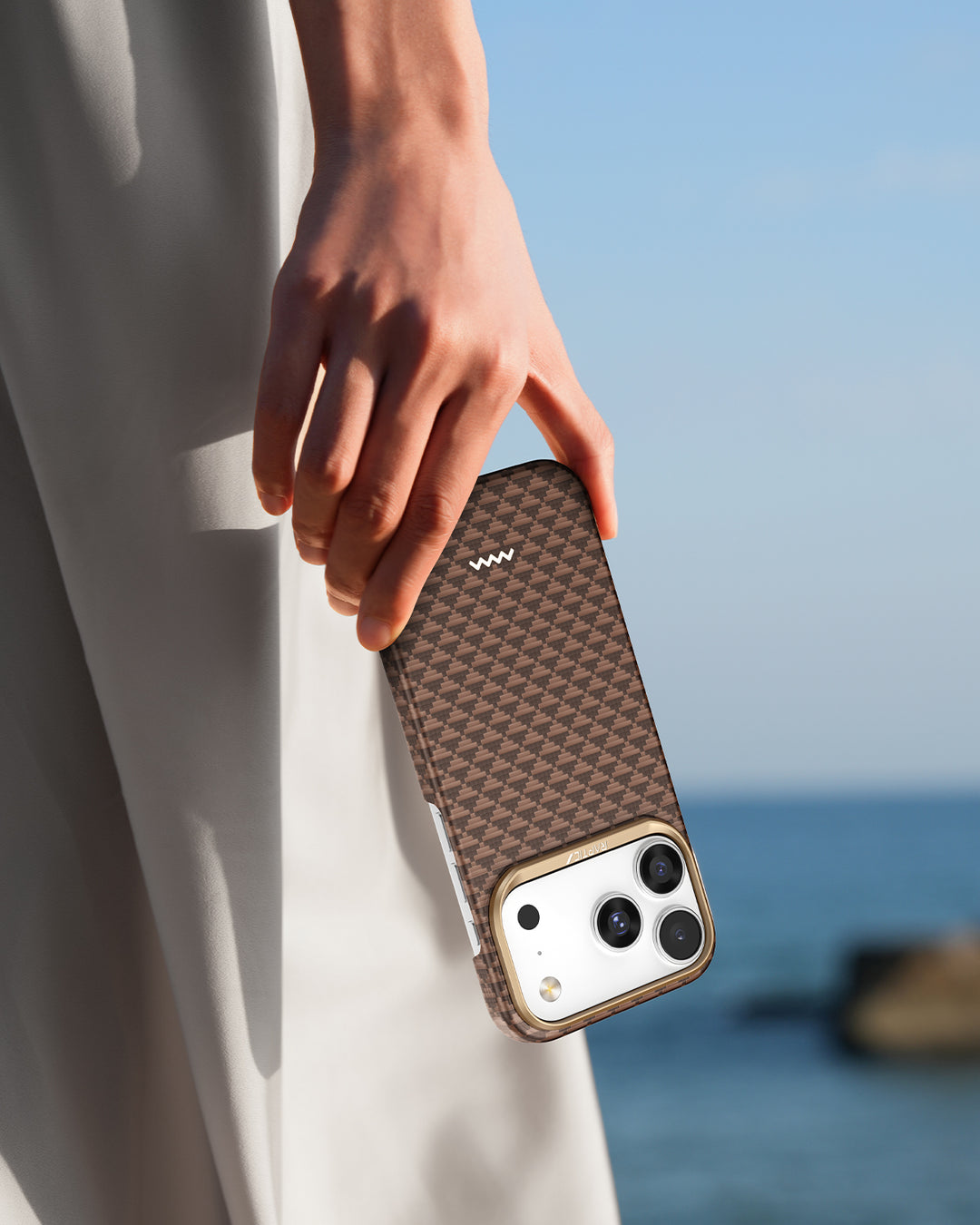 A hand holds an iPhone 17 with the Raptic Skin-Origin aramid fibre case in a brown checkered pattern. Outdoors, the soft-focus sky and water, plus a hint of white clothing, create a relaxed coastal vibe.