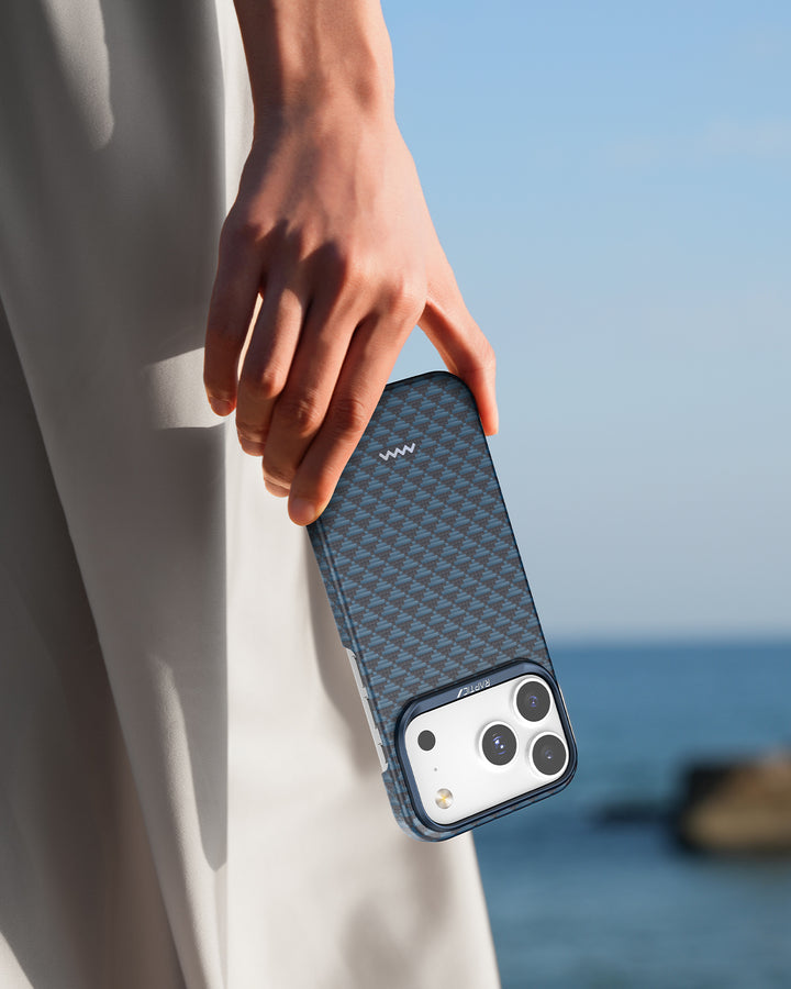 A hand holds the Raptic Skin-Origin iPhone 17 Aramid Fibre Case, featuring a blue patterned design, beside light gray pants. The clear sky and blurred water background highlight its MagSafe compatible style and ultra-slim look.
