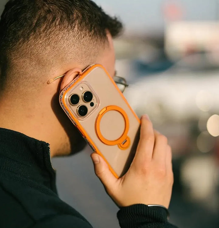 A short-haired individual holds the Raptic AirJoy Beads with Magsafe for iPhone 16 Pro Max to their ear, flaunting its clear case with air bumpers and an orange ring. Sporting clear-framed glasses and a dark jacket against a sunny blurred backdrop, they exude a modern vibe.