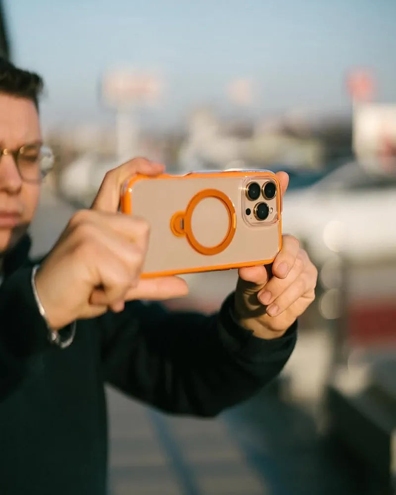 A person with glasses holds an iPhone 16 Pro Max in a Raptic AirJoy Beads with Magsafe orange case, aiming the camera outward. The blurred background shows a bright, sunny day with hints of parked cars and a blue sky.