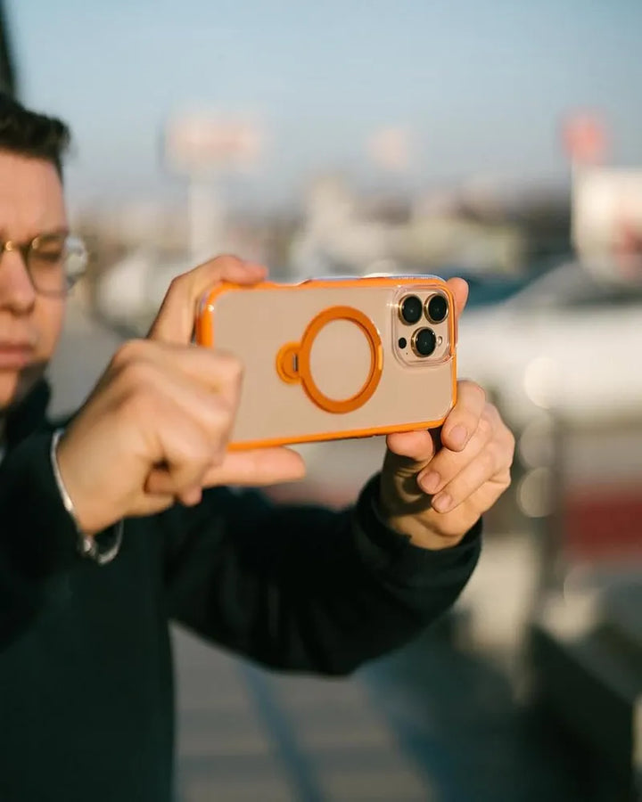 A person with glasses holds an iPhone 16 Pro Max in a Raptic AirJoy Beads with Magsafe orange case, aiming the camera outward. The blurred background shows a bright, sunny day with hints of parked cars and a blue sky.