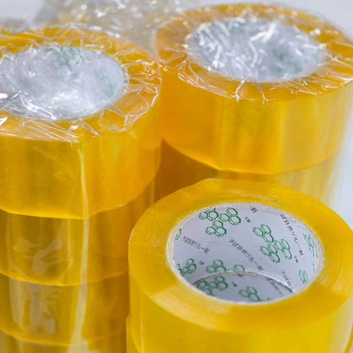 A close-up reveals vertically stacked rolls of MobileTechnica's Clear Packaging Tape with strong adhesion and tear resistance, each wrapped in transparent plastic. The inner core features a green pattern and text on a white background, giving the glossy tape a bulk packaging feel.