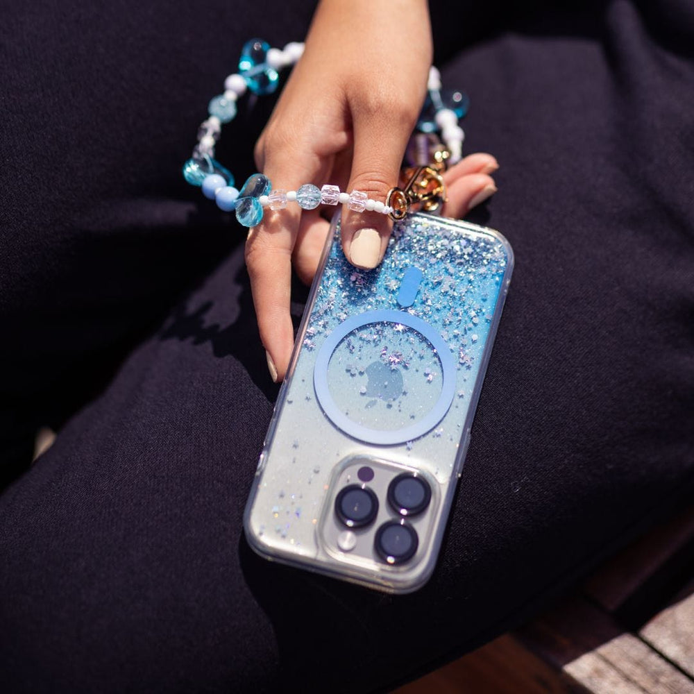 A hand holds a Bryten Starburst MagSafe case for the iPhone 16 Pro Max, featuring a glittery blue gradient design. The attached wrist strap is adorned with beads in various shades of blue, white, and gold details. The person is wearing dark pants and is seated in an outdoor setting with sunlight casting shadows.