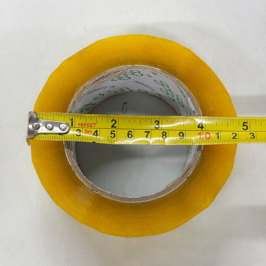 A roll of clear packaging tape by MobileTechnica lies flat on a white surface, with a yellow measuring tape stretched across its diameter, showing about 4 inches or 10 centimeters. The visible cardboard core highlights its strong adhesion and tear-resistant qualities.