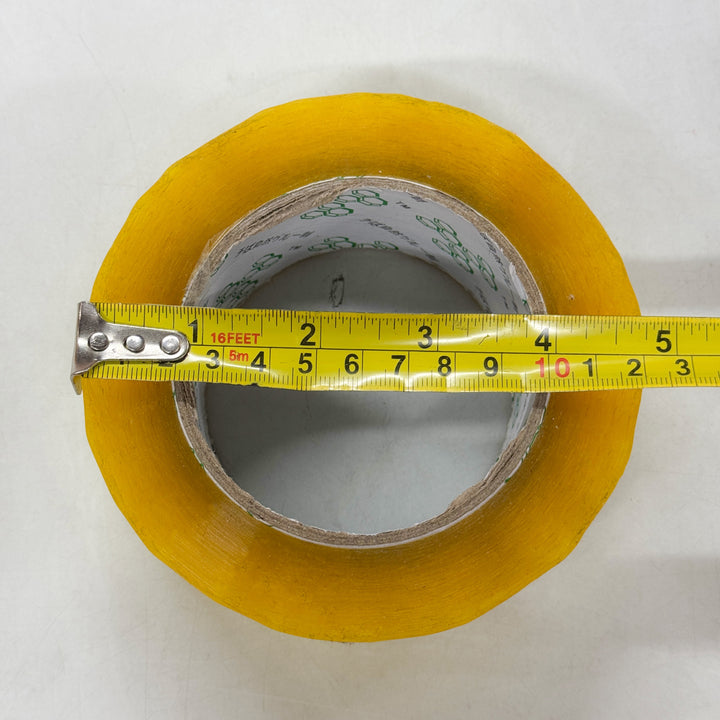 A roll of clear packaging tape by MobileTechnica lies flat on a white surface, with a yellow measuring tape stretched across its diameter, showing about 4 inches or 10 centimeters. The visible cardboard core highlights its strong adhesion and tear-resistant qualities.